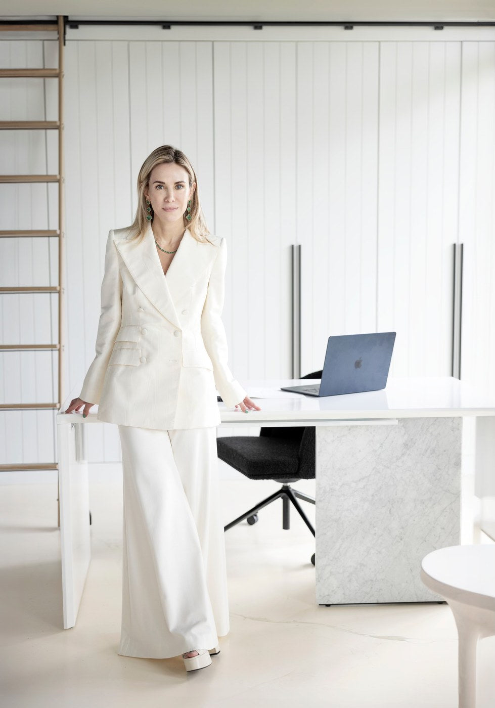 Dani Beneschott in her LVD Spaces studio — a cream double-breasted suit, malachite earrings, against vertical white shiplap and a Carrara marble desk.