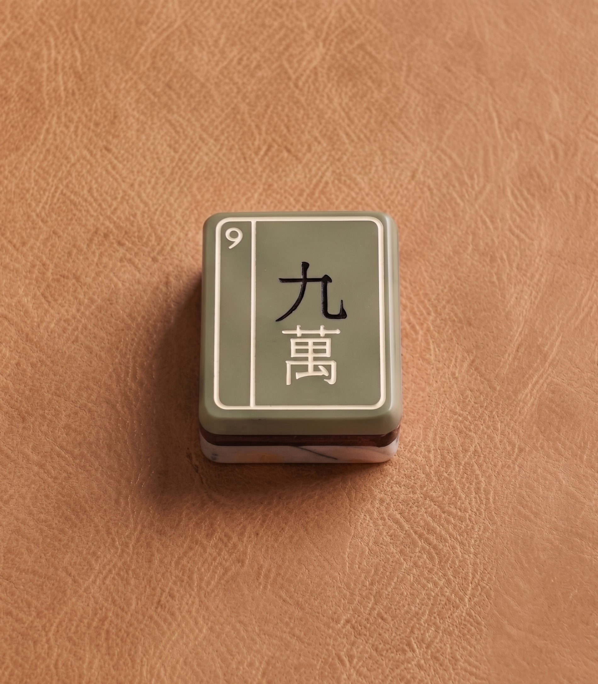 Three-layer acrylic Mise en Mahjong tile shown at scale — sage face, black core, ivory pearl acrylic back with rose-gold mark.